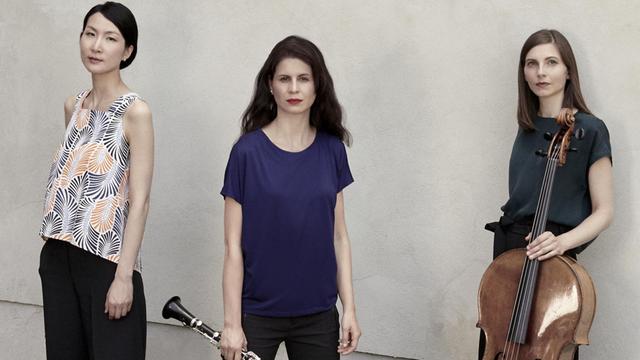 Drei Frauen stehen vor einer betonierten Wand und schauen mit neutralem Ausdruck in die Kamera. Die Frau in der Mitte hat eine Klarinette in der Hand und die Frau rechts ein Cello.