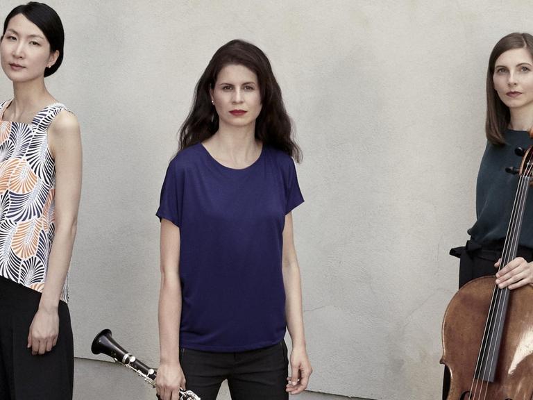 Drei Frauen stehen vor einer betonierten Wand und schauen mit neutralem Ausdruck in die Kamera. Die Frau in der Mitte hat eine Klarinette in der Hand und die Frau rechts ein Cello.