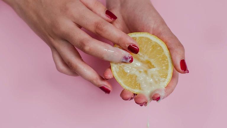 Vor rosa Hintergrund halten zwei Hände mit rotem Nagellack eine Zitronenhälfte, über die eine milchige Flüssigkeit läuft.