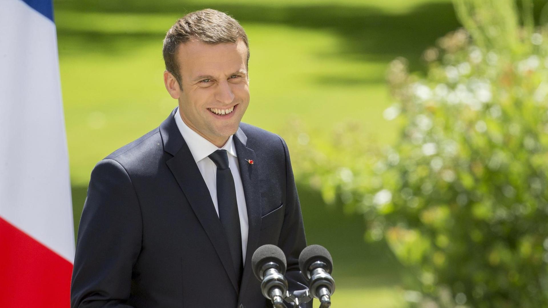 Emmanuel Macron steht neben einer französischen Flagge und lacht. Emmanuel Macron steht neben einer französischen Flagge und lacht.