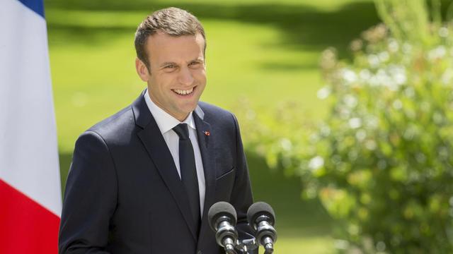 Emmanuel Macron steht neben einer französischen Flagge und lacht.