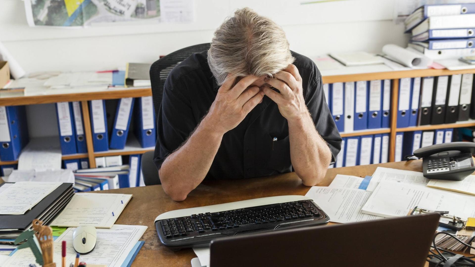 Ein Mann sitzt an einem Büroarbeitsplatz und hält seine Hände vor das Gesicht. Die Ellbogen sind dabei auf dem Schreibtisch abgestützt. Auf dem Schreibtisch befinden sich ein PC, ein Telefon und Unterlagen. Im Hintergrund sind Regale mit Büroordnern darin Ein Mann sitzt an einem Büroarbeitsplatz und hält seine Hände vor das Gesicht. Die Ellbogen sind dabei auf dem Schreibtisch abgestützt. Auf dem Schreibtisch befinden sich ein PC, ein Telefon und Unterlagen. Im Hintergrund sind Regale mit Büroordnern darin