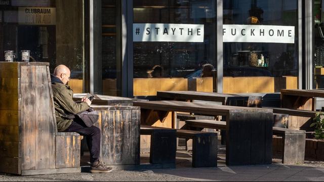 Fassade eines Restaurants, in dessen Schaufenster der Aufruf "Stay The Fuck At Home" zu sehen ist. Auf der Sitzgelegenheit davor sitzt ein einsamer Mann.
