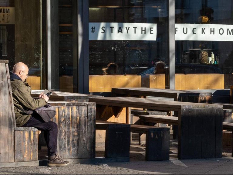 Fassade eines Restaurants, in dessen Schaufenster der Aufruf "Stay The Fuck At Home" zu sehen ist. Auf der Sitzgelegenheit davor sitzt ein einsamer Mann.