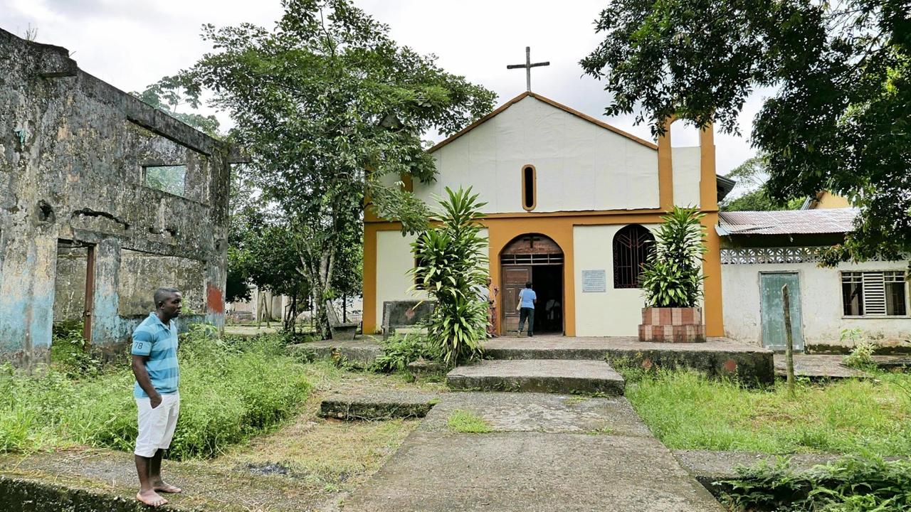 Eine kleine Dorfkirche mit Kreuz an der Spitze. Daneben die Ruinen von einem Gebäude und ein ehemaliger afrokolumbianischer Dorfbewohner.