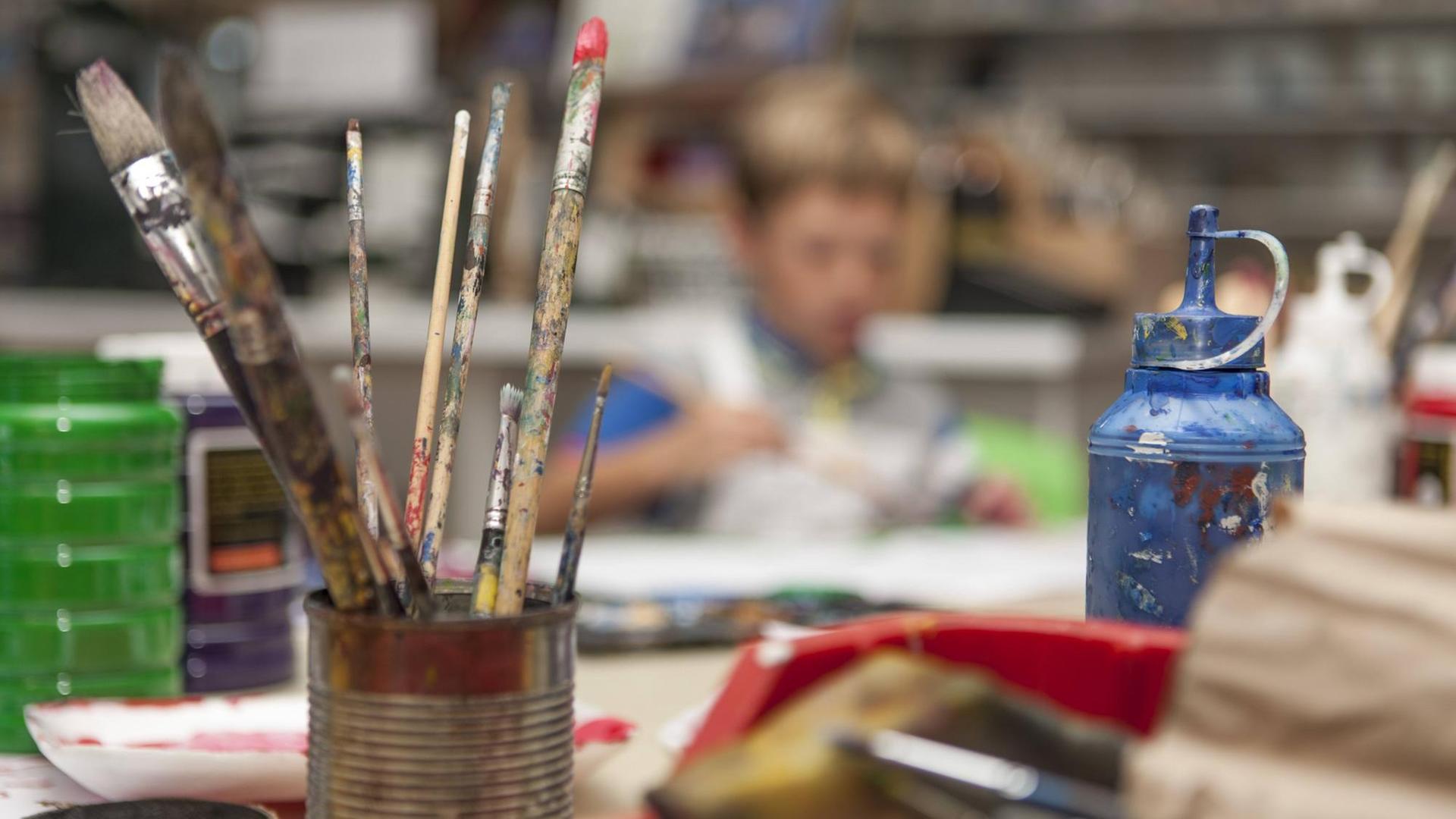 Farben und Pinsel stehen auf einem Tisch, im Hintergrund sitzen Kinder. Farben und Pinsel stehen auf einem Tisch, im Hintergrund sitzen Kinder.