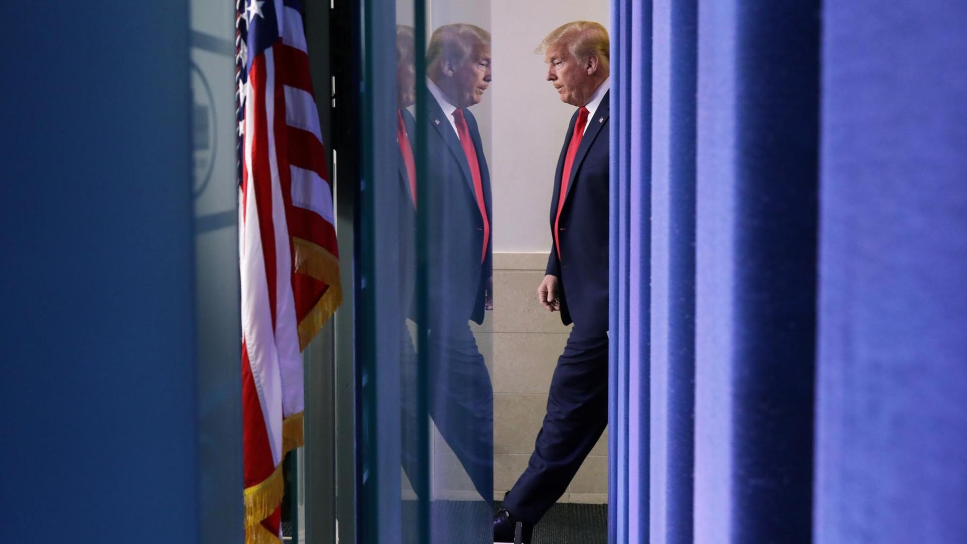 Profilansicht von Trump mit Spieglung in einer Scheibe. Links im Bild die amerikanische Flagge.