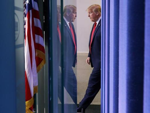 Profilansicht von Trump mit Spieglung in einer Scheibe. Links im Bild die amerikanische Flagge.