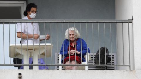 Auf einem Balkon eines Pflegeheims sitzt eine ältere Dame, Neben ihr steht eine Pflegerin mit Mundschutz.