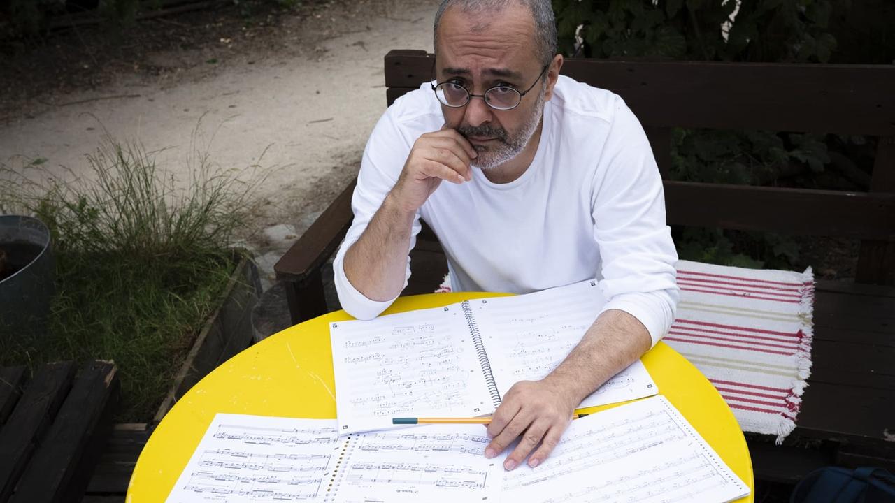 Ein Mann mit Brille sitzt an einem gelben, runden Gartentisch voller Notenblätter. Ein Mann mit Brille sitzt an einem gelben, runden Gartentisch voller Notenblätter.