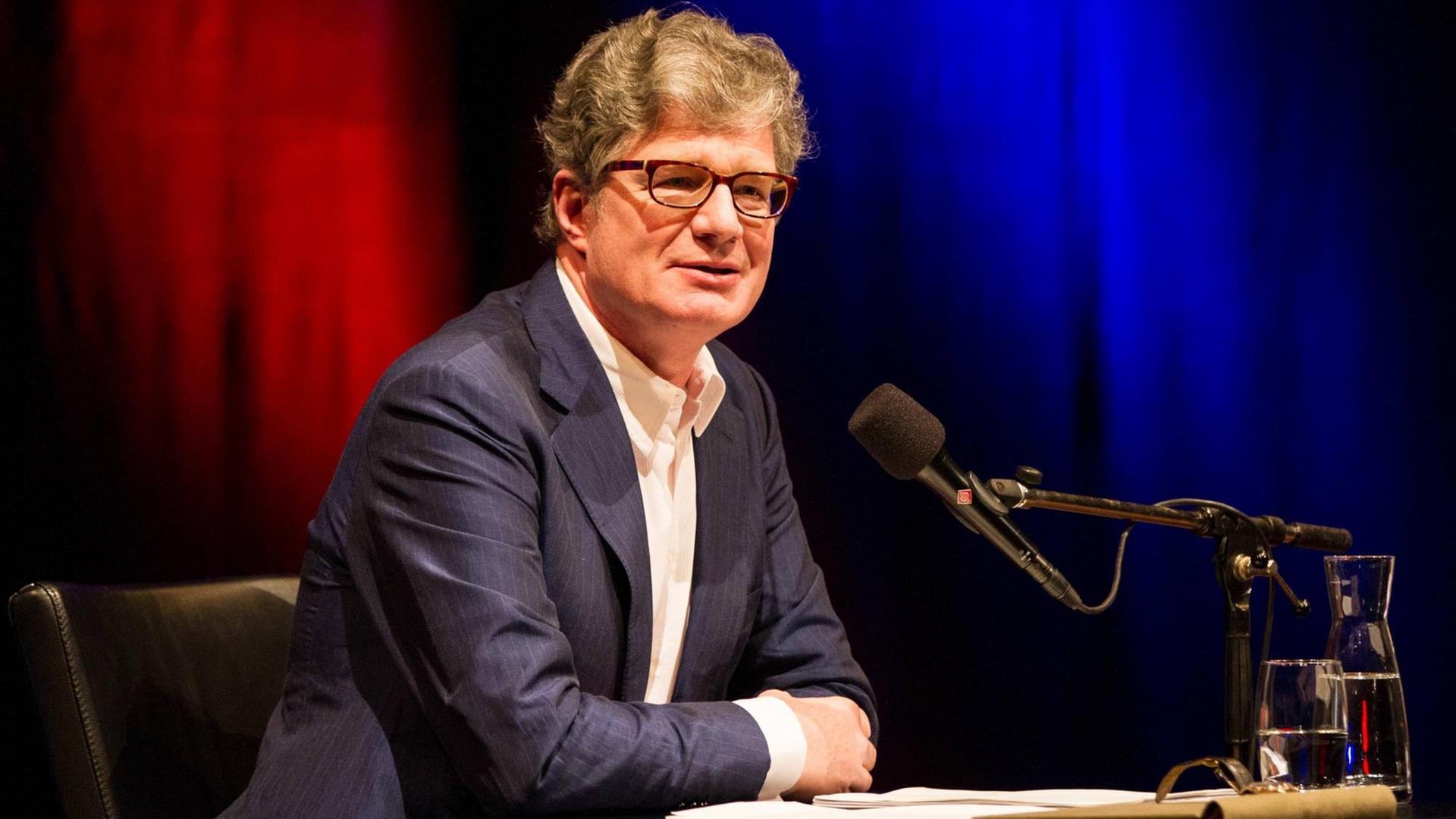 Roger Willemsen sitzt vor einem rotblauem Hintergrund am Mikrofon