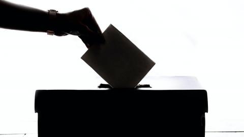 Eine Hand steckt einen Umschlag in eine Wahlurne.
