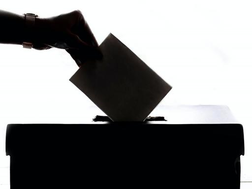 Eine Hand steckt einen Umschlag in eine Wahlurne.