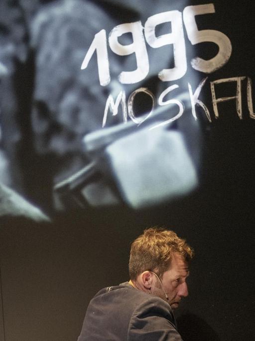Szenenausschnitt aus "Die Katze und der General" von Nino Haratischwili in der REGIE von Jette Steckel, in dem André Szymanski eine schwarze Bühnenwand mit weißer Farbe beschreibt. Im Hintergrund eine Einblendung von russischen Soldaten und darüber steht geschrieben: "1995 Moskau".