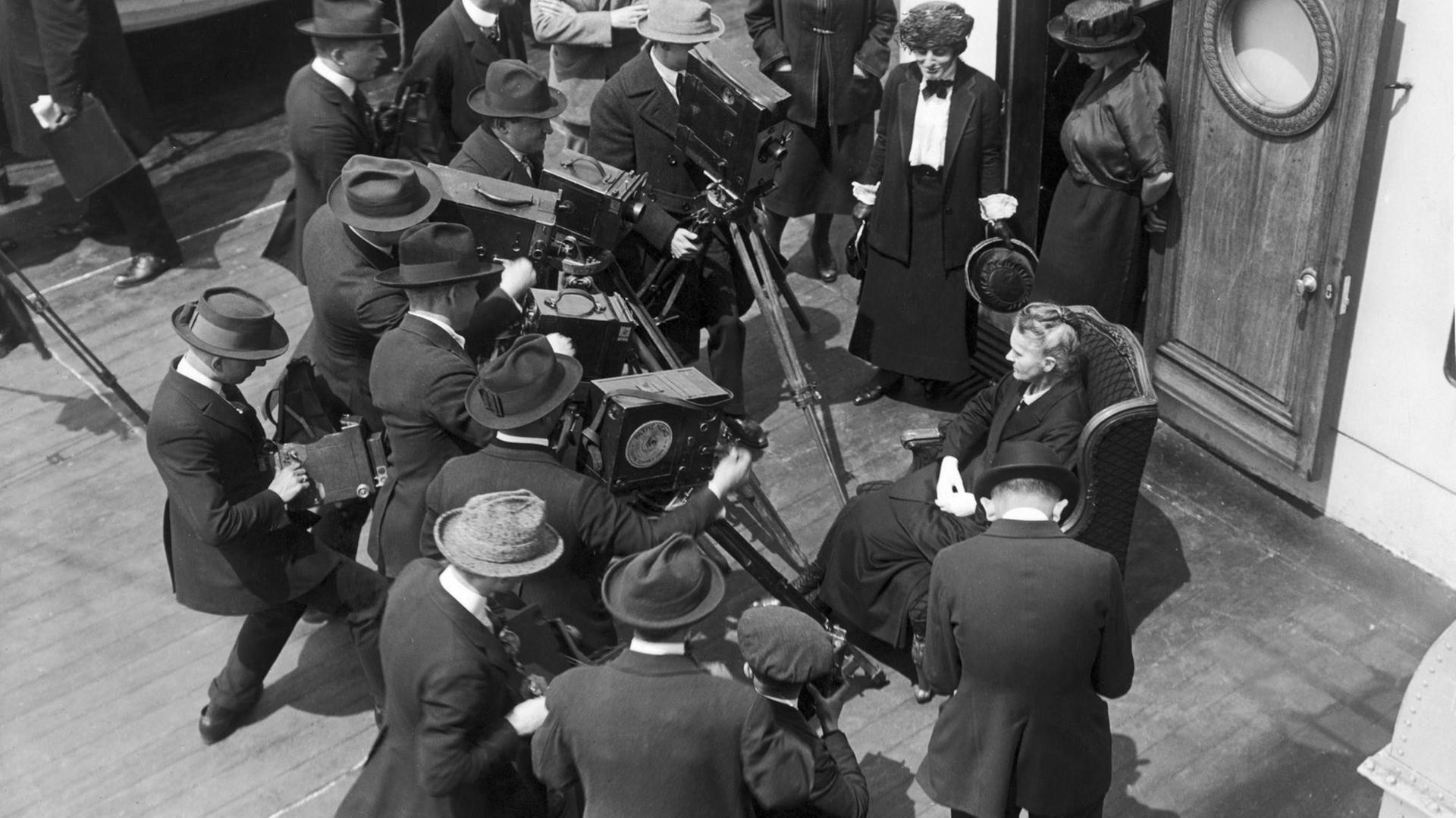 Marie Curie auf einem Schiff, umgeben von Kameramännern. Marie Curie auf einem Schiff, umgeben von Kameramännern.