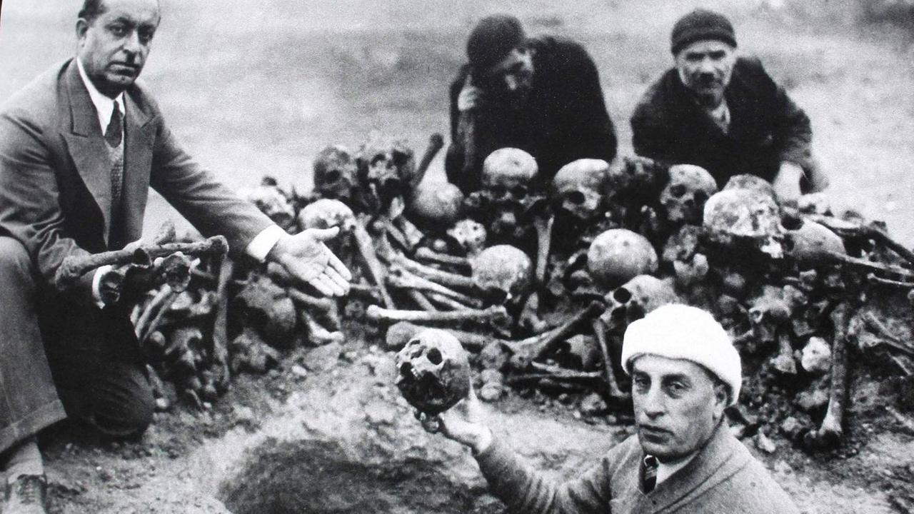Historisches Foto von Armeniern vor einem Haufen Schädel und Gebeinen. Ein Mann hält einen Totenschädel hoch. 
