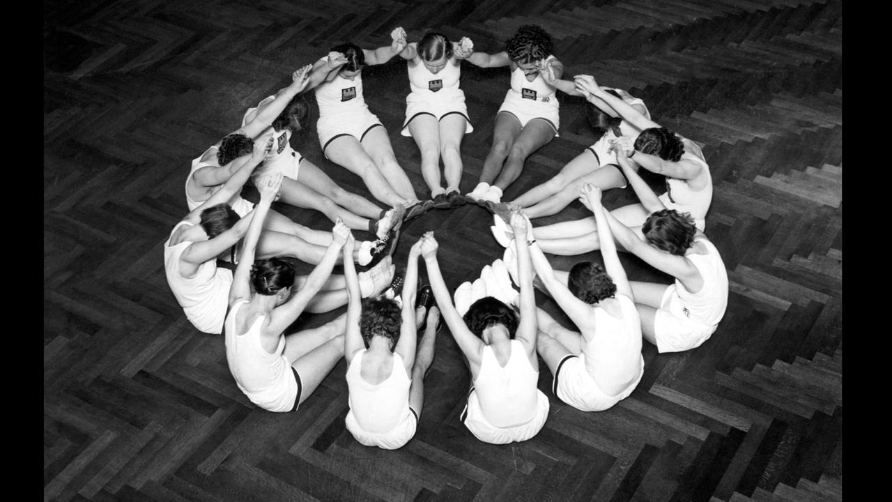 13 Frauen in kurzer weißer Sportkleidung sitzen auf dem Boden im Kreis. Sie haben ihre Beine zur Mitte des Kriese gestreckt, ihre Arme in die Höhe gehoben und den Kopf nach vorn gebeugt. 13 Frauen in kurzer weißer Sportkleidung sitzen auf dem Boden im Kreis. Sie haben ihre Beine zur Mitte des Kriese gestreckt, ihre Arme in die Höhe gehoben und den Kopf nach vorn gebeugt.