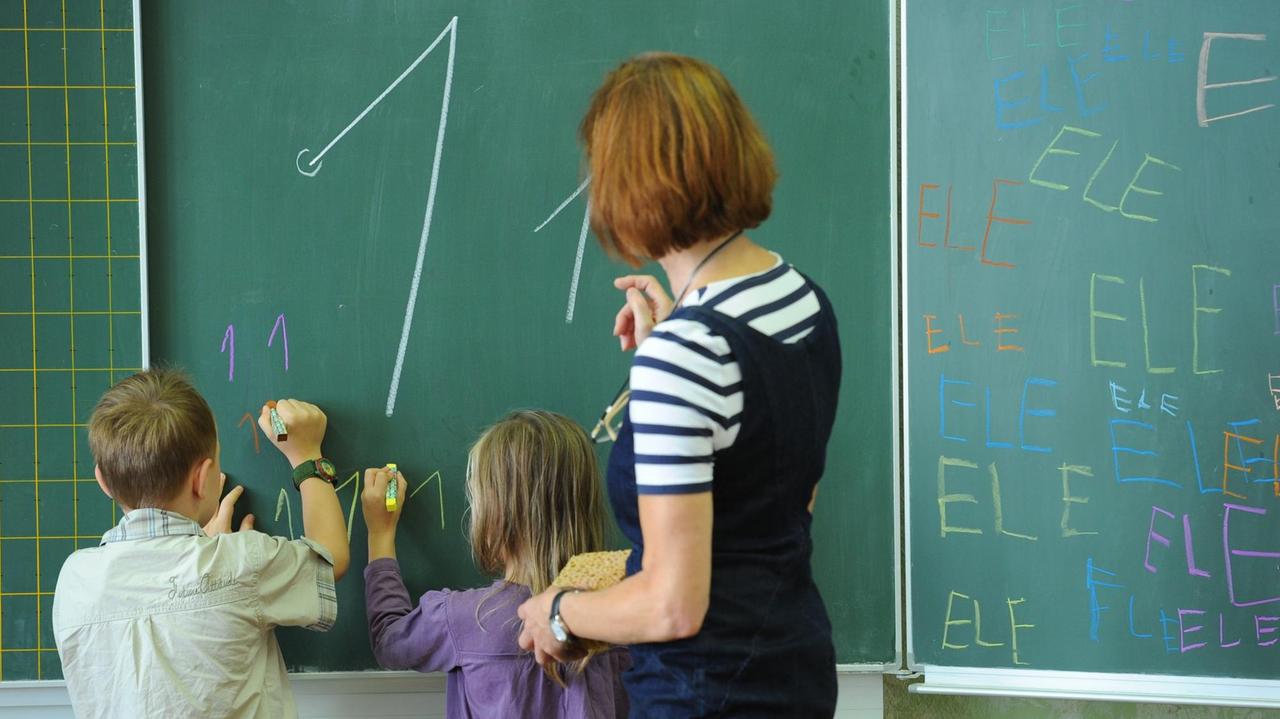 Eine Lehrerin steht mit einer Schülerin und einem Schüler an einer Tafel und schreibt eine 1. Eine Lehrerin steht mit einer Schülerin und einem Schüler an einer Tafel und schreibt eine 1.