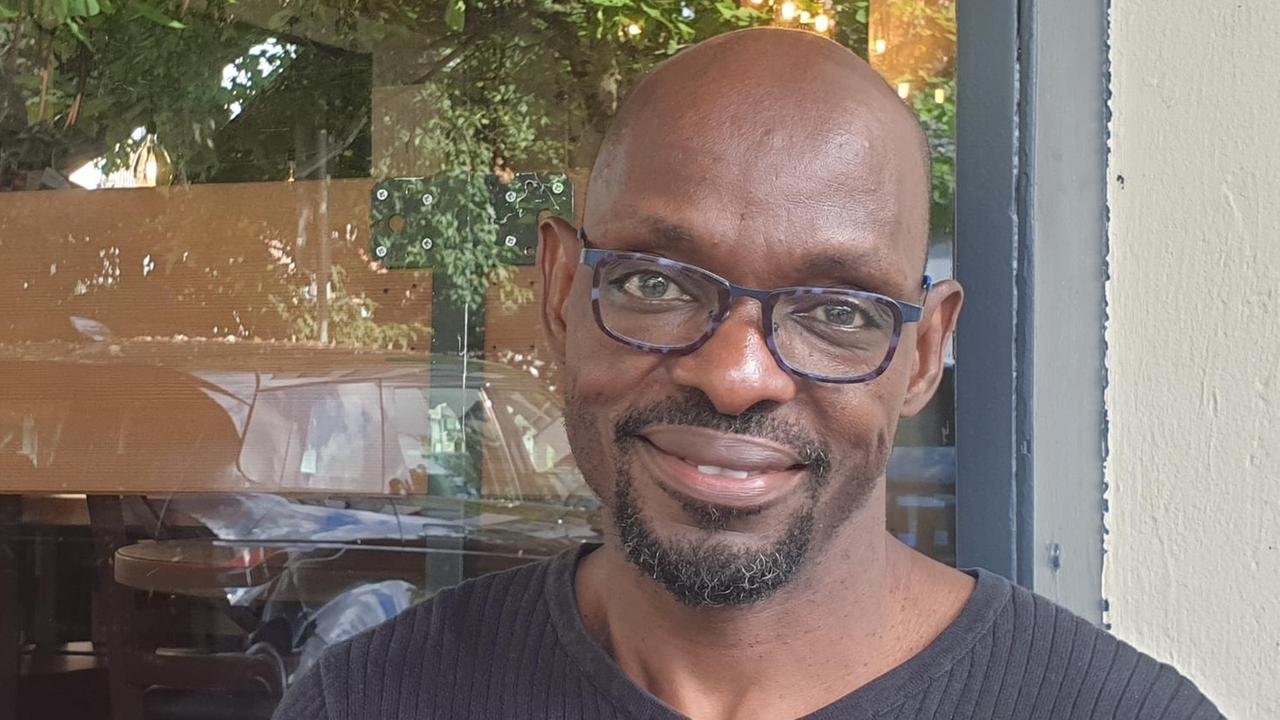 Abdoulaye Sounaye, mit Brille in einem schwarzen Pullover, sitzt im freien vor einer weiß gekalkten Hauswand, im Fenster hinter ihm spiegeln sich Autos und grünes Blattwerk. Abdoulaye Sounaye, mit Brille in einem schwarzen Pullover, sitzt im freien vor einer weiß gekalkten Hauswand, im Fenster hinter ihm spiegeln sich Autos und grünes Blattwerk.