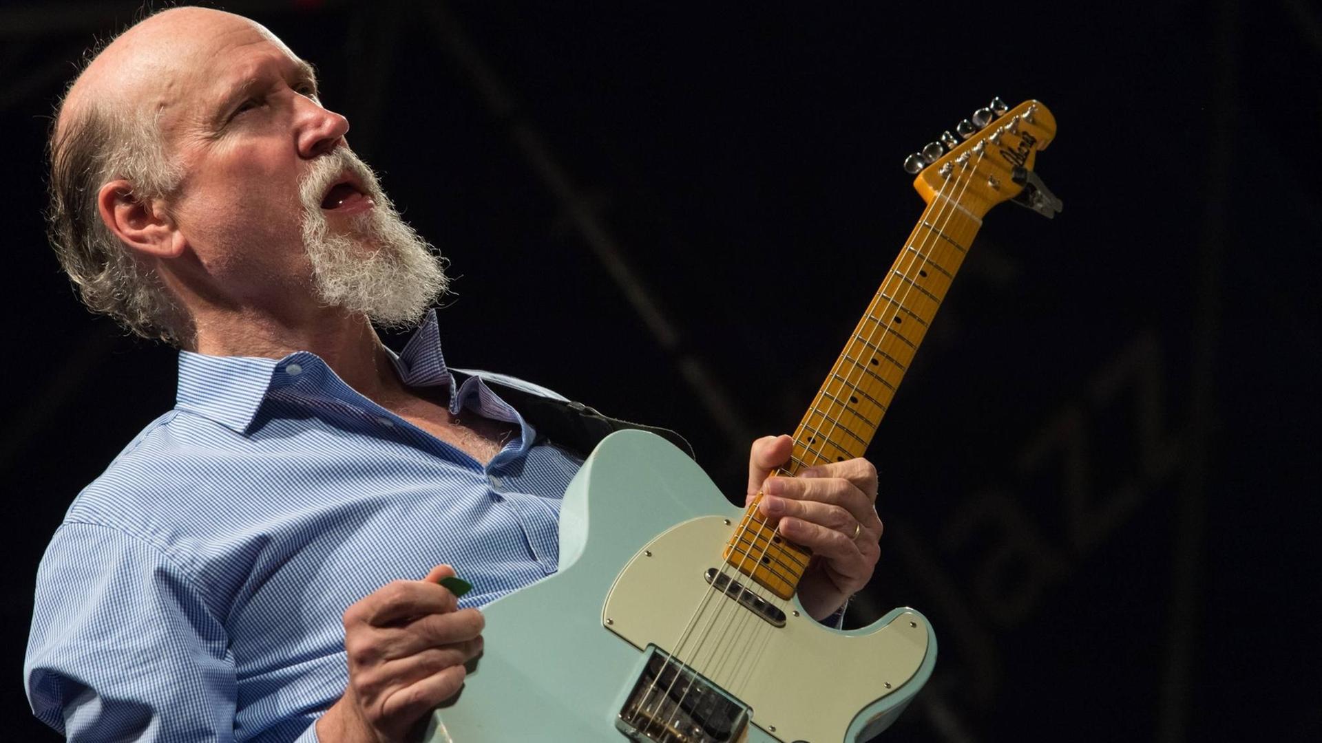 Ein bärtiger älterer Mann im blauen Hemd hält eine mintgrüne E-Gitarre in der Hand. Ein bärtiger älterer Mann im blauen Hemd hält eine mintgrüne E-Gitarre in der Hand.