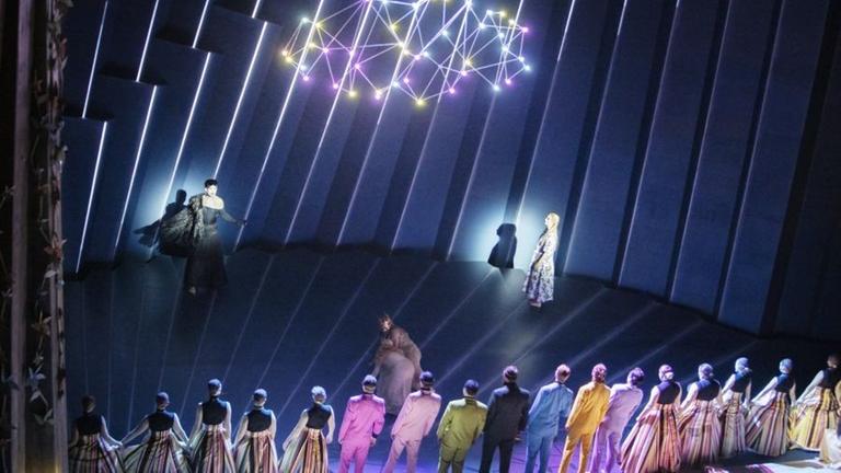 Im Vordergrund mehrere bunt gekleidete Menschen, die eine Mauer bilden, dahinter zwei in Gold gekleidete Frauen mit Kronen auf ihren Häupter, die sich gegenseitig stützen, noch weiter hinten eine schwarz gekleidete Frau und eine Frau in einem Kleid mit Blumenmuster. Im Vordergrund mehrere bunt gekleidete Menschen, die eine Mauer bilden, dahinter zwei in Gold gekleidete Frauen mit Kronen auf ihren Häupter, die sich gegenseitig stützen, noch weiter hinten eine schwarz gekleidete Frau und eine Frau in einem Kleid mit Blumenmuster.