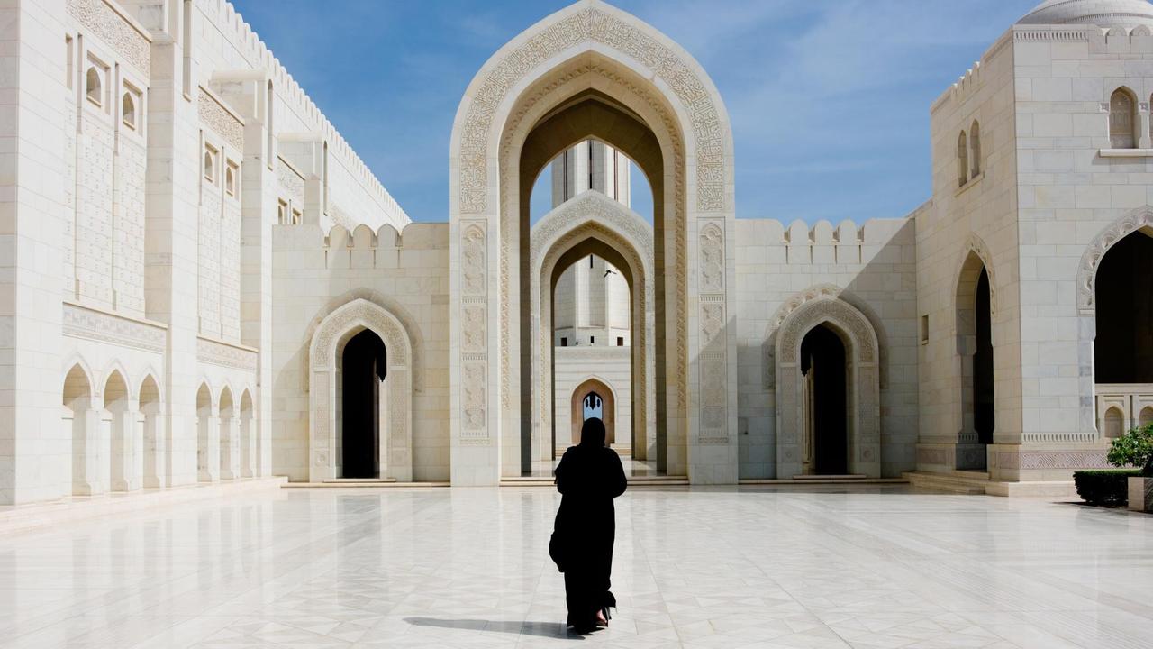 Frau in schwarzem Abaya-Kleid läuft  auf den Bogen in der berühmten Großen Moschee des Sultans Qaboos in Maskat zu.