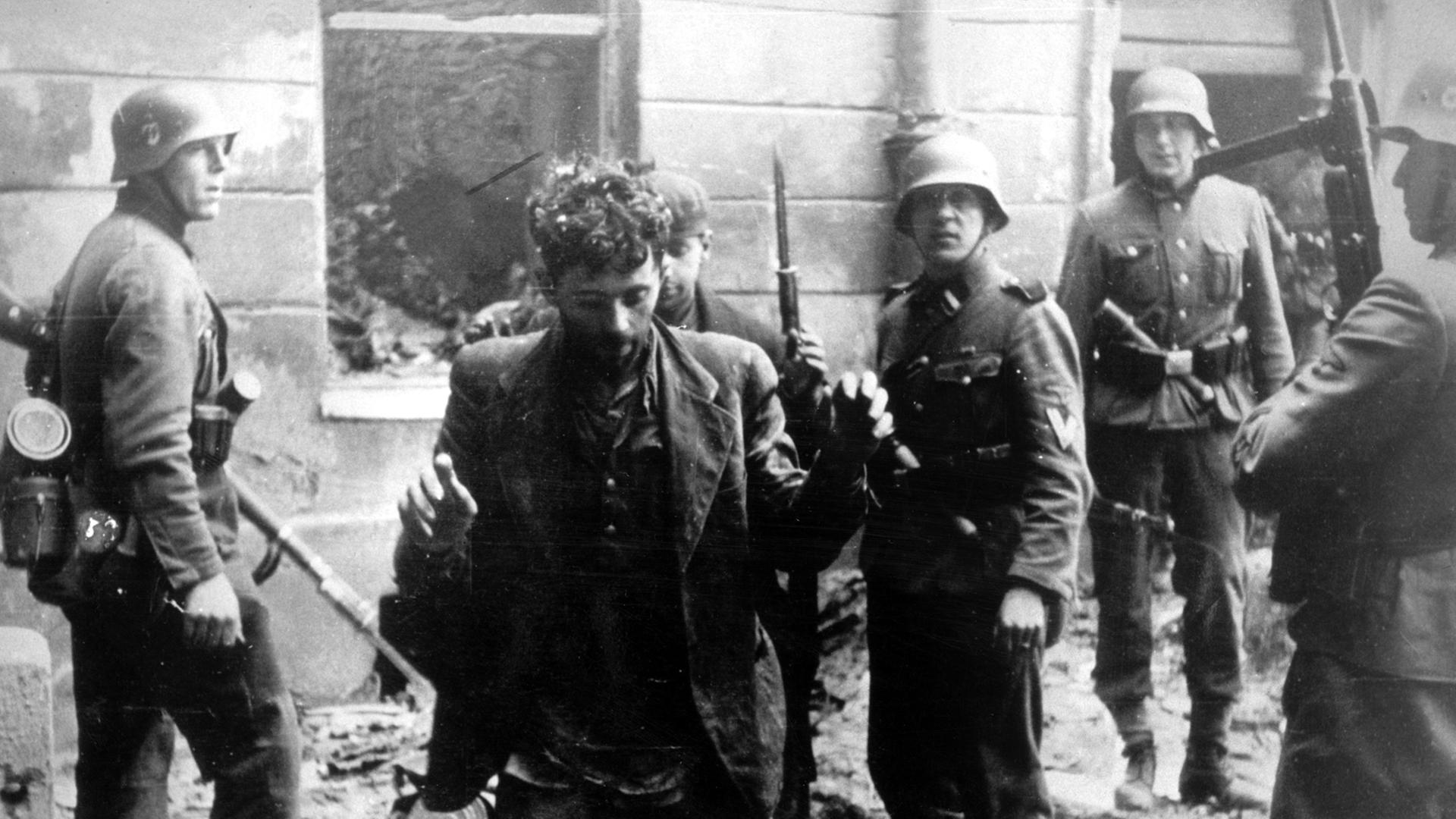 Zwei Juden, die sich in einem Haus versteckt hatten, werden von SS-Soldaten gefangen genommen. Die Aufnahme entstand während des Warschauer Ghetto-Aufstands, der vom 19. April 1943 bis zu seiner blutigen Niederschlagung am 16. Mai 1943 dauerte. Die Nationalsozialisten hatten ein Jahr nach der Besetzung Polens im November 1940 in Warschau ein Ghetto errichtet und dorthin annähernd eine halbe Million Juden verschleppt. Zwischen Juli und September 1942 wurden 300 000 Opfer in den Todeslagern, die meisten in Treblinka, ermordet. Als am 19. April 1943 die SS mit der Verschleppung der restlichen 60 000 Ghetto-Einwohner begann, leisteten mehrere hundert militärisch organisierte Juden bewaffneten Widerstand. Bei den Kämpfen wurden etwa 14 000 jüdische Aufständische getötet. Zwei Juden, die sich in einem Haus versteckt hatten, werden von SS-Soldaten gefangen genommen. Die Aufnahme entstand während des Warschauer Ghetto-Aufstands, der vom 19. April 1943 bis zu seiner blutigen Niederschlagung am 16. Mai 1943 dauerte. Die Nationalsozialisten hatten ein Jahr nach der Besetzung Polens im November 1940 in Warschau ein Ghetto errichtet und dorthin annähernd eine halbe Million Juden verschleppt. Zwischen Juli und September 1942 wurden 300 000 Opfer in den Todeslagern, die meisten in Treblinka, ermordet. Als am 19. April 1943 die SS mit der Verschleppung der restlichen 60 000 Ghetto-Einwohner begann, leisteten mehrere hundert militärisch organisierte Juden bewaffneten Widerstand. Bei den Kämpfen wurden etwa 14 000 jüdische Aufständische getötet.