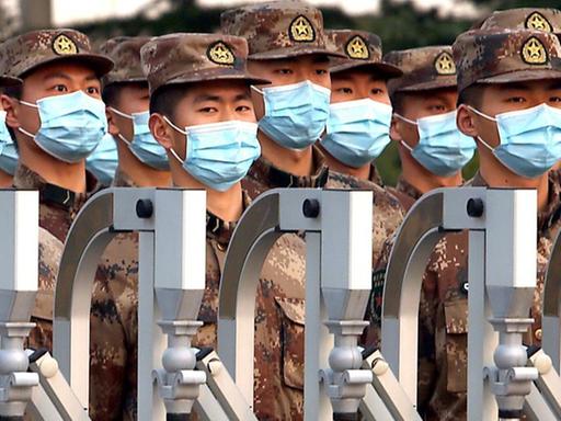Chinesische Soldaten mit OP-Masken maschieren hinter einem Zaun.