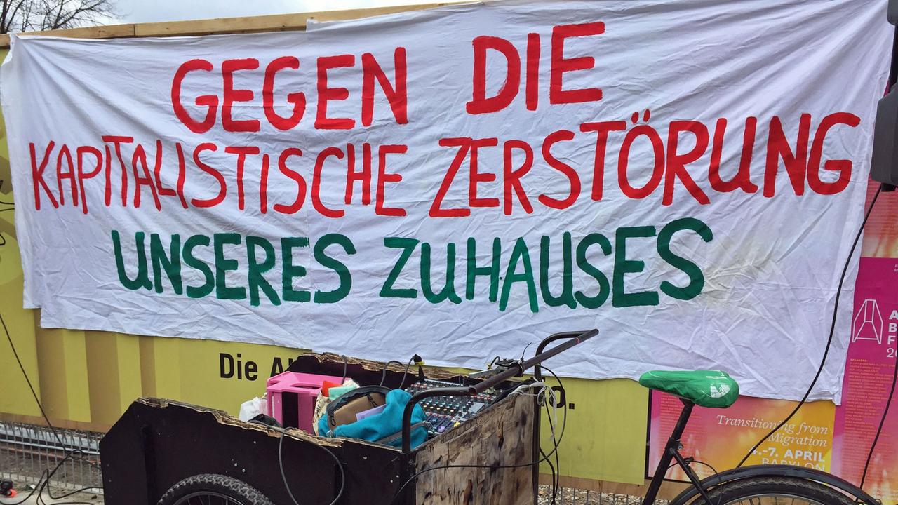 An einem Bauzaun hängt ein Transparent, darauf ist mit bunter Farbe geschrieben: "Gegen die kapitalistische Zerstörung unseres Zuhauses", davor steht ein Lastenrad.