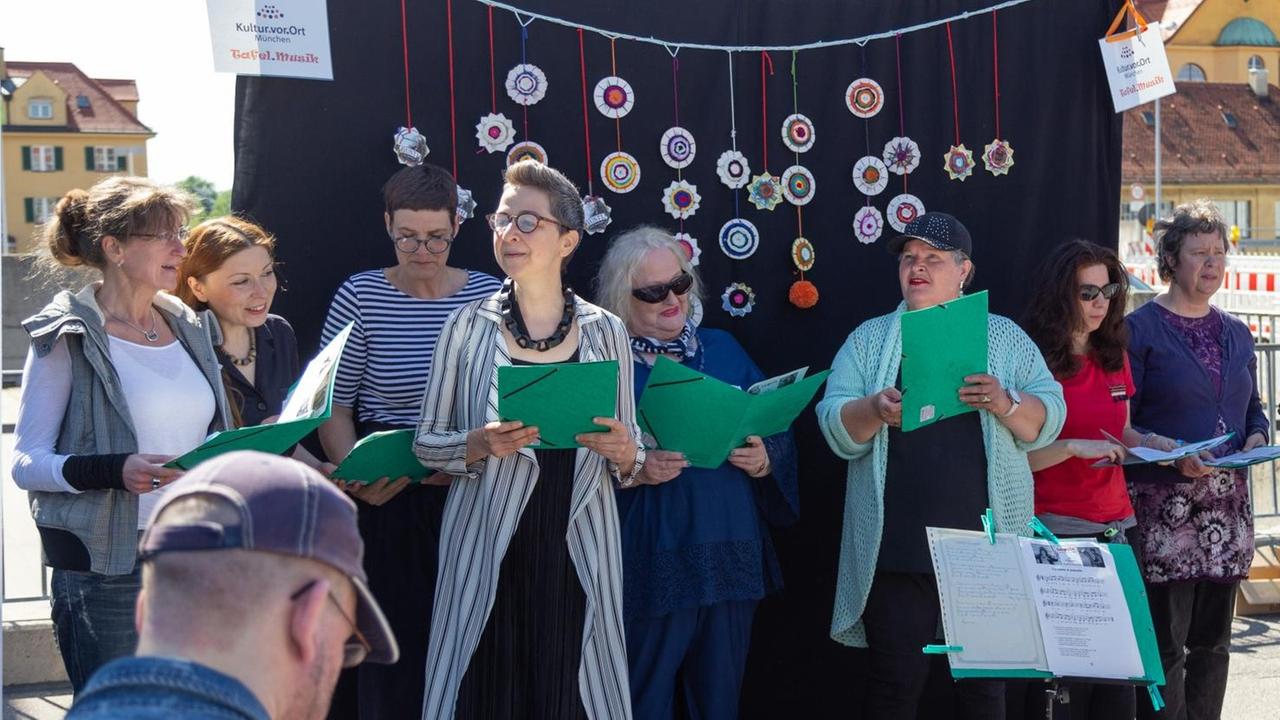 Auf einer provisorisch aufgebauten Bühne stehen Sängerinnen, die aus grünen Chormappen singen. Auf einer provisorisch aufgebauten Bühne stehen Sängerinnen, die aus grünen Chormappen singen.