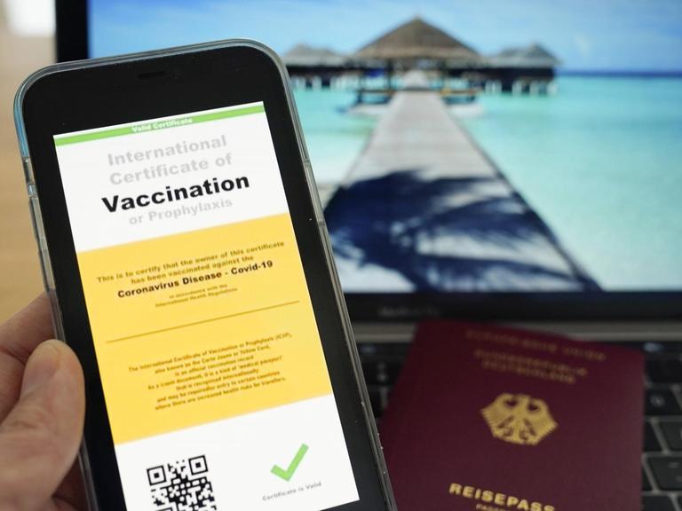 Symbolfoto: Digitaler Impfass und Reisepass vor Fernweh-Motiv auf einem Laptop.