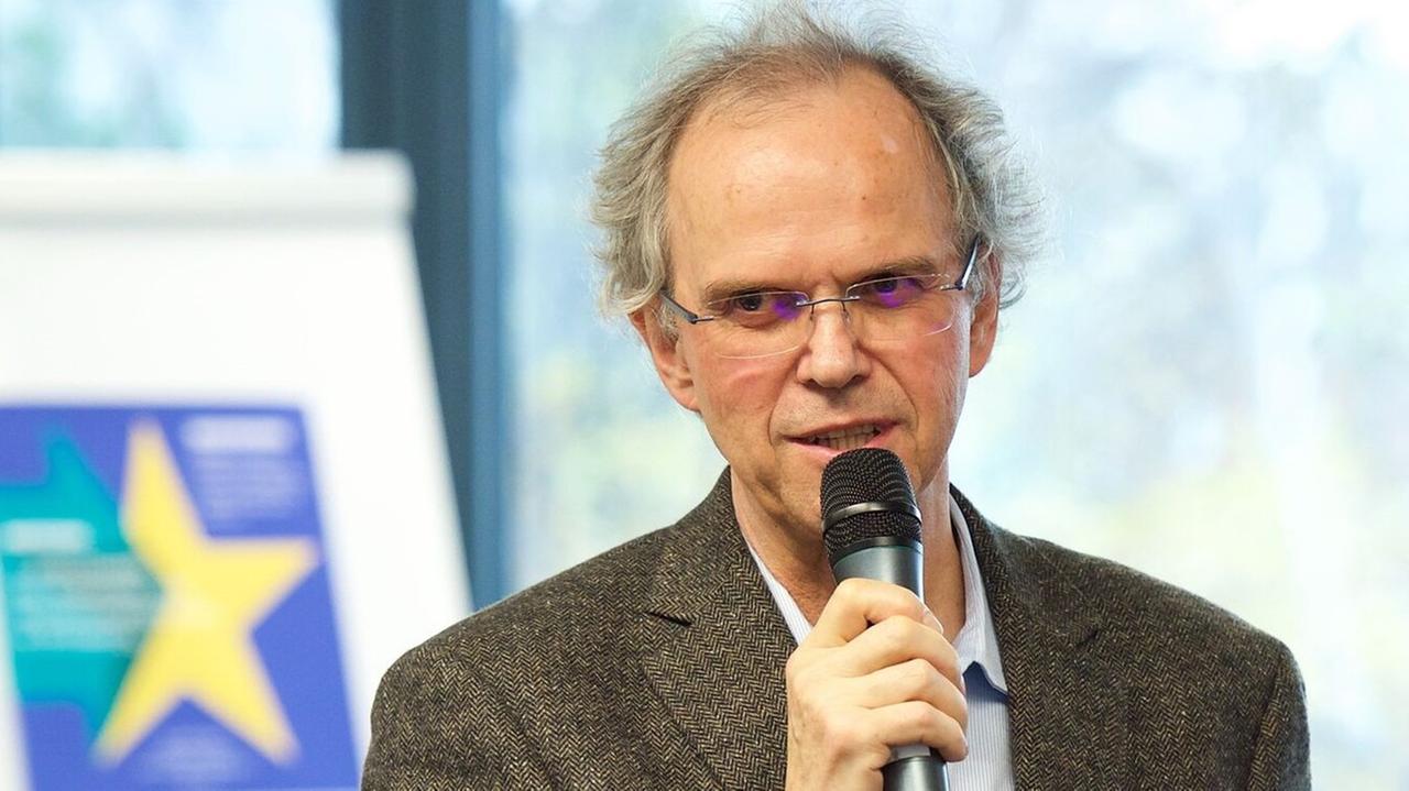Rainer Bauböck, mit Brille im grauen Sakko, steht in einem Seminarraum und spricht in ein Mikrofon, das er in der rechten Hand hält.