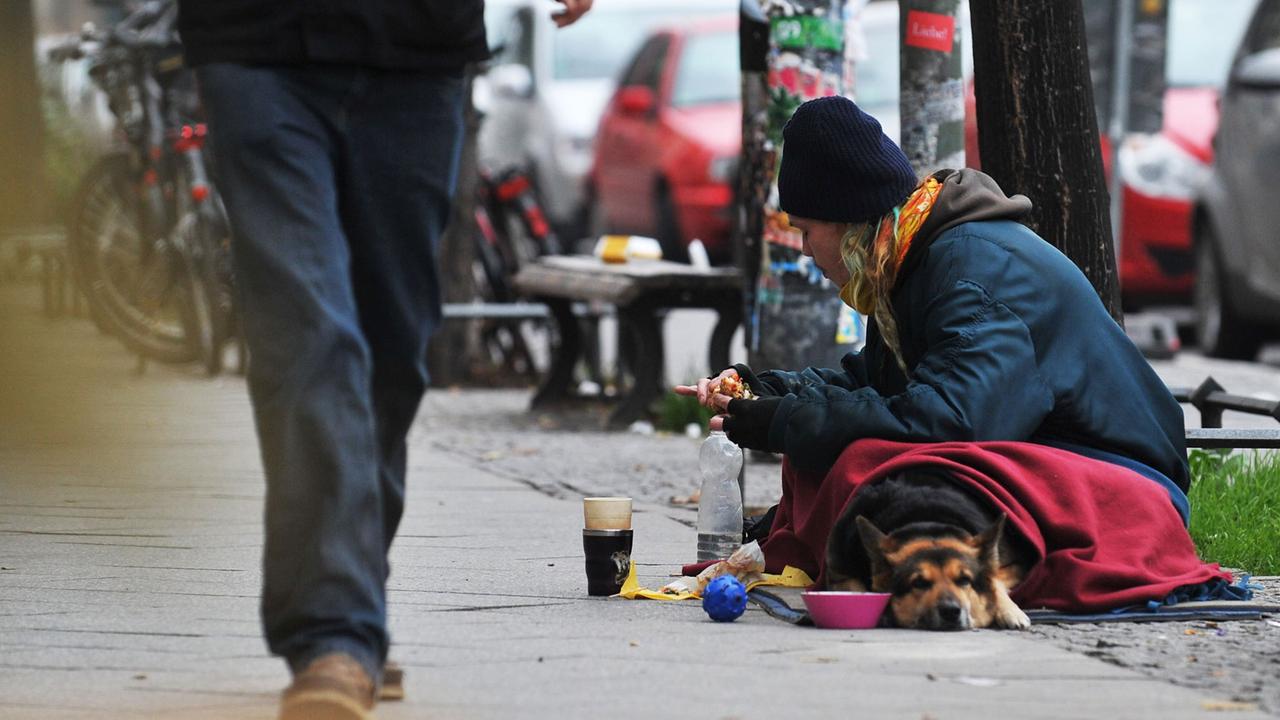 Auf einem Gehweg sitzt ein Mann in einer Decke. Vor ihm steht ein Becher. Ein Passant geht vorbei. Auf einem Gehweg sitzt ein Mann in einer Decke. Vor ihm steht ein Becher. Ein Passant geht vorbei.