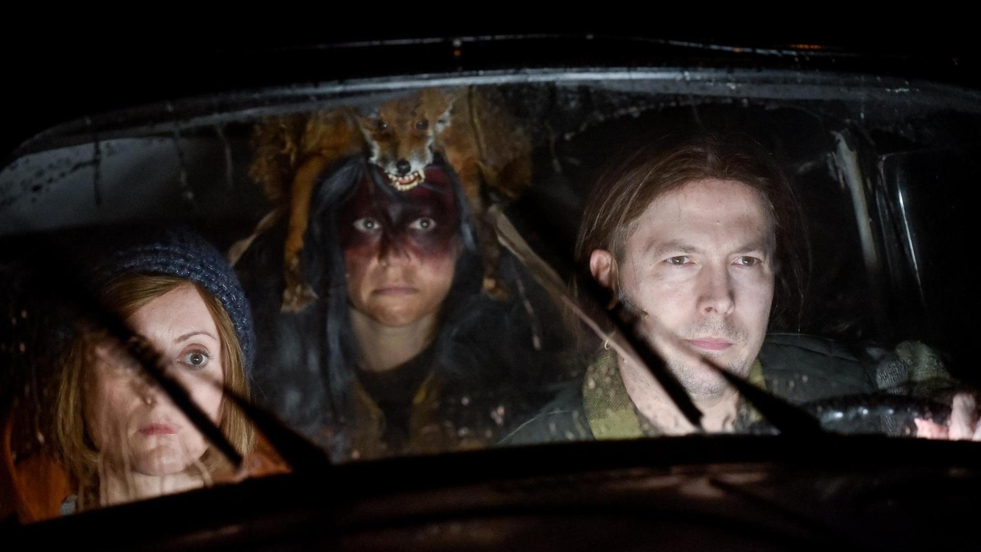 Der Szenenausschnitt aus dem Stück "Die Anderen" von Anne-Cécile Vandalem an der Berliner Schaubühne zeigt durch die Frontscheibe eines Autos ins Innere blickend Veronika Bachfischer, Ruth Rosenfeld (als Schamanin verkleidet), David Ruland im Auto sitzend mit in der Bewegung erstarrten Scheibenwischern. Der Szenenausschnitt aus dem Stück "Die Anderen" von Anne-Cécile Vandalem an der Berliner Schaubühne zeigt durch die Frontscheibe eines Autos ins Innere blickend Veronika Bachfischer, Ruth Rosenfeld (als Schamanin verkleidet), David Ruland im Auto sitzend mit in der Bewegung erstarrten Scheibenwischern.