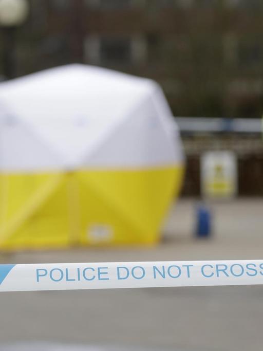 Die Polizisten in leuchtgelben Warnwesten sind unscharf im Vordergrund zu sehen. Im Hintergrund ein gelb-weißes Zelt, das über den Tatort gespannt wurde und das von blau-weißem Absperrband umgeben ist.