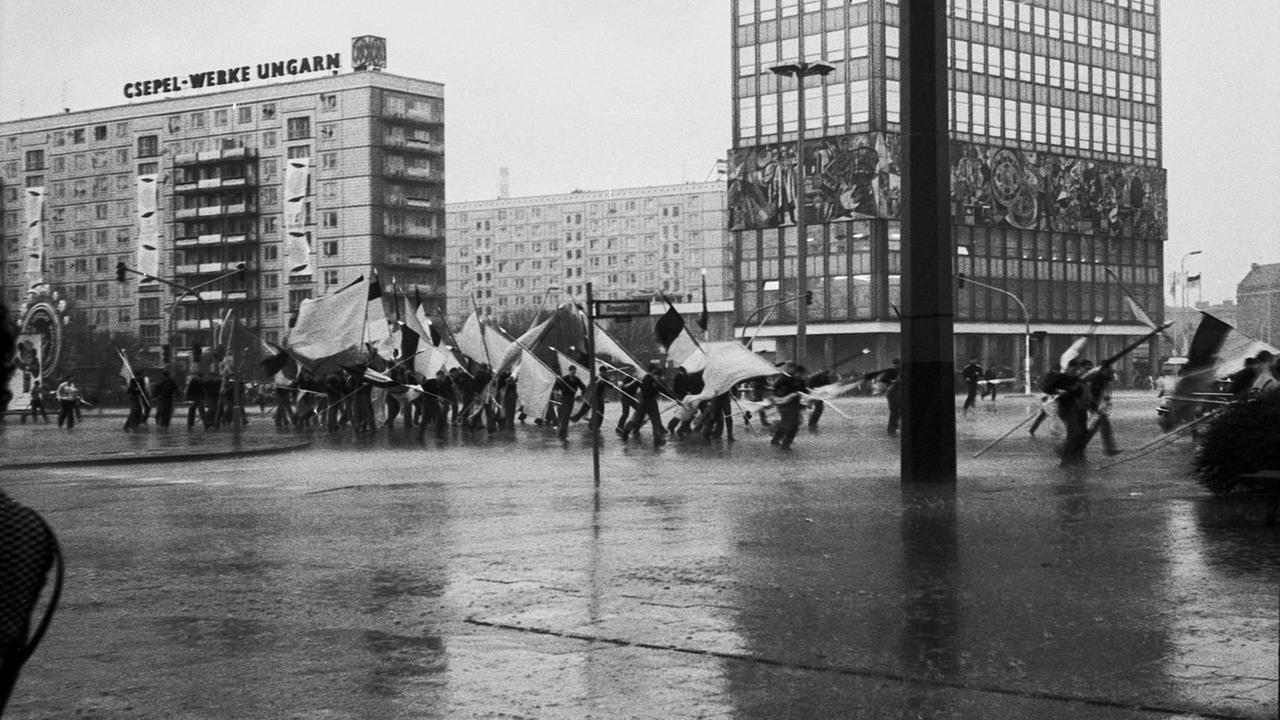 Offizielles Foto der Podiumsdiskussion im Hau: Ein Demonstrationszug läuft mit Fahnen auf den Straßen zwischen Plattenbauten entlang.