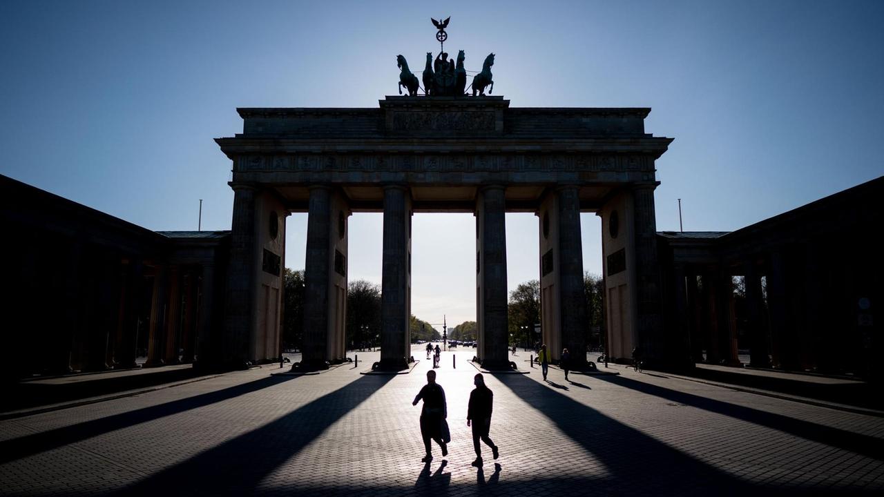 Das Brandenburger Tor in Berlin wirft in der untergehenden Sonne lange Schatten, zwei Personen gehen mittig davor entlang.