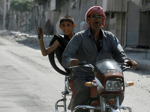 Ein Mann und ein Junge fahren auf einem Motorroller durch eine Straße in Manbidsch, der Junge winkt in die Kamera.