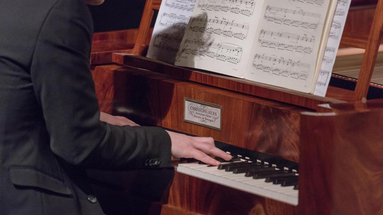 An einem holzfarbenen und -gemaserten hammerflügel sieht man die Hände eines Mannes in schwarzem Anzug auf der Klaviatur liegen. Ein Schild am Instrument zeigt den Namen des Klavierbauers, auf dem Notenpult stehen aufgeschlagene Noten. An einem holzfarbenen und -gemaserten hammerflügel sieht man die Hände eines Mannes in schwarzem Anzug auf der Klaviatur liegen. Ein Schild am Instrument zeigt den Namen des Klavierbauers, auf dem Notenpult stehen aufgeschlagene Noten.