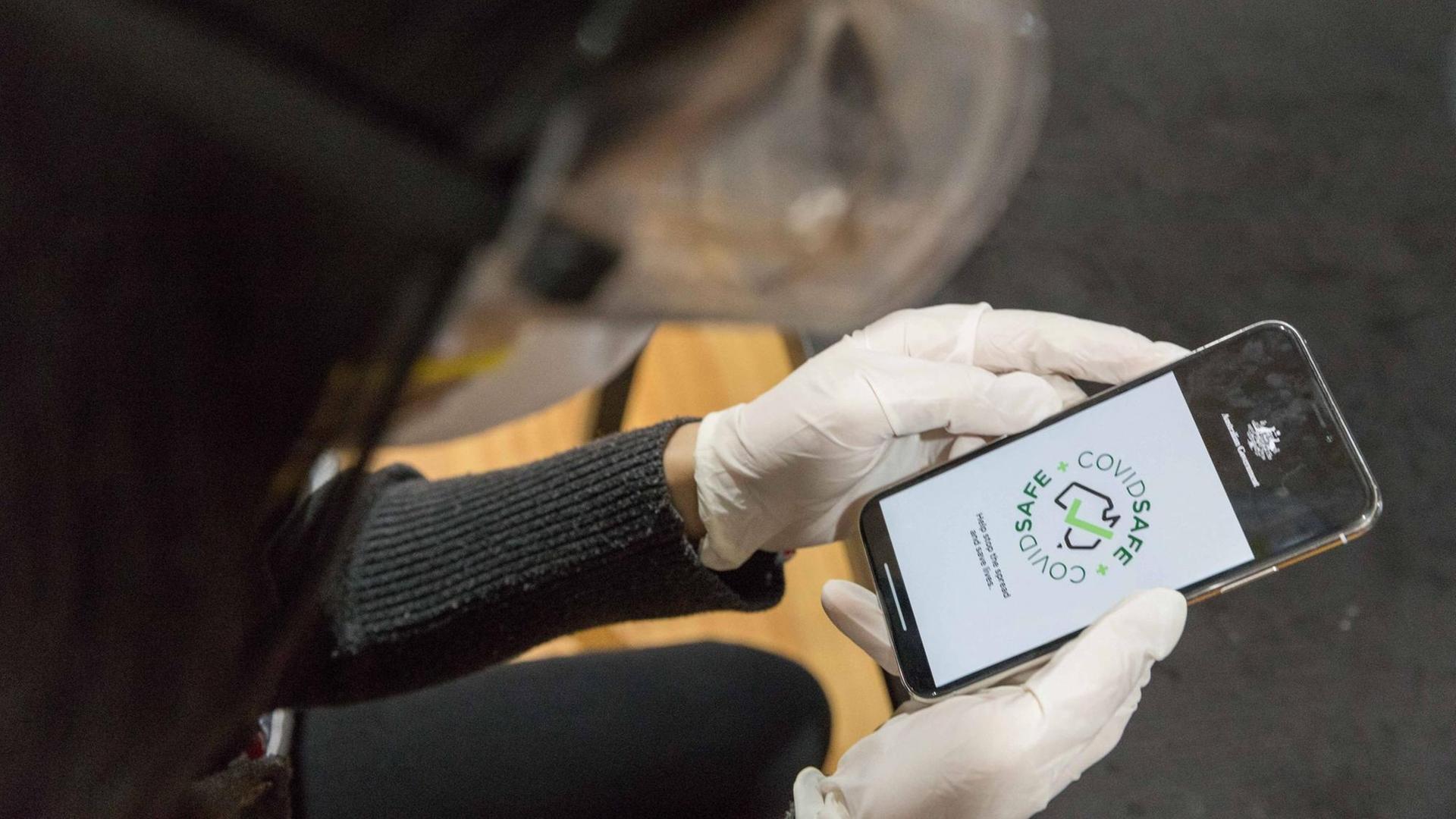 Hände in Plastik-Einweghandschuhen halten ein Smartphone, auf dessen Display die Corona-App "CovidSafe" angezeigt wird. Hände in Plastik-Einweghandschuhen halten ein Smartphone, auf dessen Display die Corona-App "CovidSafe" angezeigt wird.