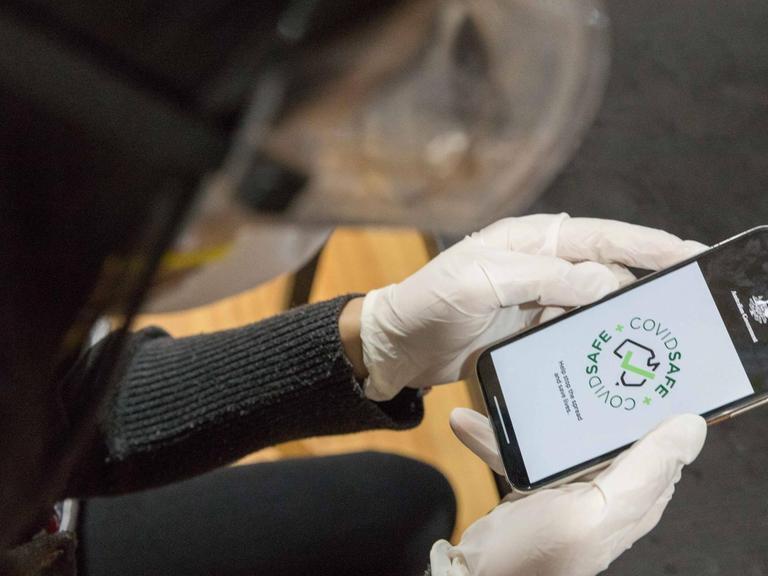 Hände in Plastik-Einweghandschuhen halten ein Smartphone, auf dessen Display die Corona-App "CovidSafe" angezeigt wird.