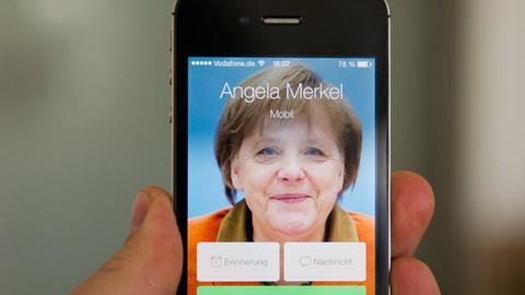 Ein Mann hält ein Apple iPhone 4S, auf dem der fiktive Kontakt Angela Merkel anruft und ein Bild der Bundeskanzlerin zu sehen ist.