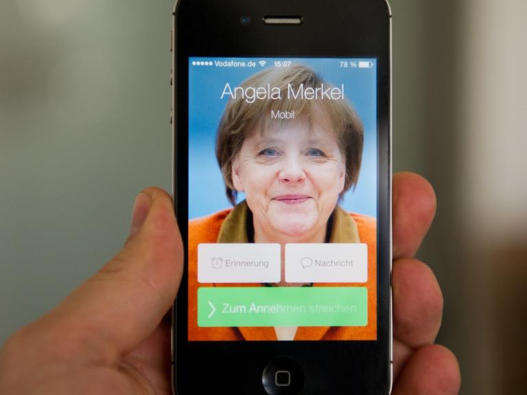 Ein Mann hält ein Apple iPhone 4S, auf dem der fiktive Kontakt Angela Merkel anruft und ein Bild der Bundeskanzlerin zu sehen ist.