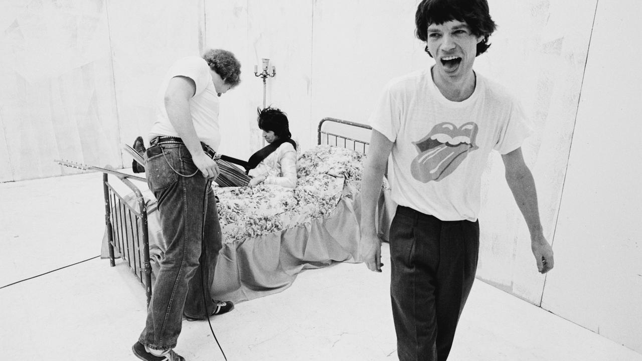 Rolling-Stones-Frontmann Mick Jagger trägt bei einer Musikvideoproduktion ein T-Shirt mit dem Bandlogo - einer herausgestreckten Zunge - und streckt selbst die Zunge heraus. Im Hintergrund liegt Gitarrist Keith Richards auf einem Bett. Rolling-Stones-Frontmann Mick Jagger trägt bei einer Musikvideoproduktion ein T-Shirt mit dem Bandlogo - einer herausgestreckten Zunge - und streckt selbst die Zunge heraus. Im Hintergrund liegt Gitarrist Keith Richards auf einem Bett.