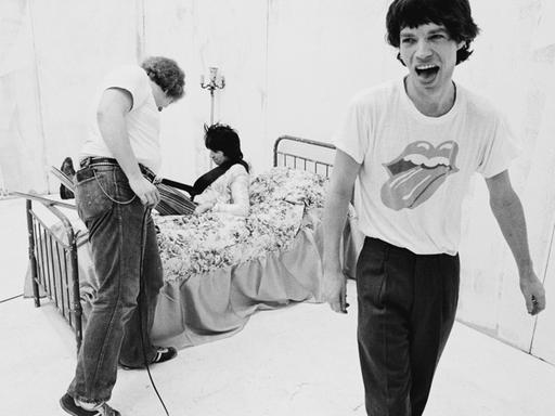 Rolling-Stones-Frontmann Mick Jagger trägt bei einer Musikvideoproduktion ein T-Shirt mit dem Bandlogo - einer herausgestreckten Zunge - und streckt selbst die Zunge heraus. Im Hintergrund liegt Gitarrist Keith Richards auf einem Bett.