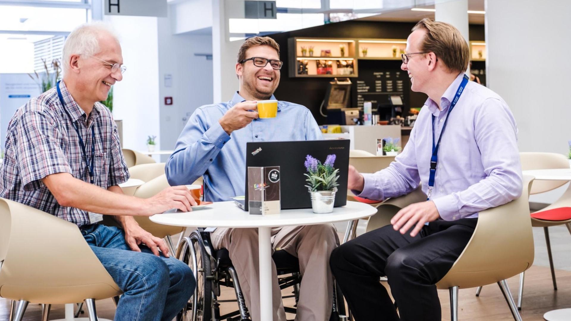 Drei Männer in Hemd und Jeans sitzen an einem runden Tisch um einen Laptop und lachen.