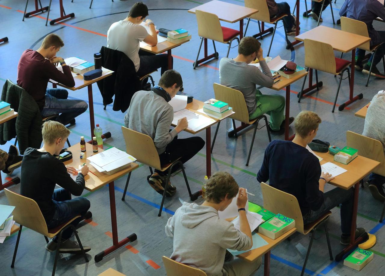 Schülerinnen und Schüler sitzen in einer Turnhalle an Einzeltischen und machen Abiturprüfungen. Vor ihnen liegen Englisch-Wörterbücher. Schülerinnen und Schüler sitzen in einer Turnhalle an Einzeltischen und machen Abiturprüfungen. Vor ihnen liegen Englisch-Wörterbücher.