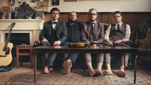 Vier Männer in Anzug und/oder Weste sitzen barfuss in einem Kaminzimmer. Vor ihnen liegt eine Mandoline auf dem Tisch.