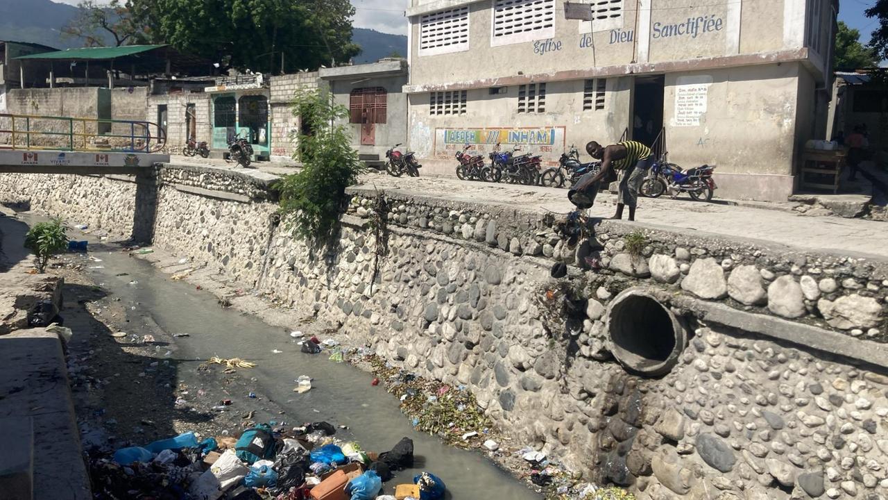 Ein Mann kippt einen Eimer mit Abfällen eine Mauer hinunter in ein kärgliches Gewässer, in dem schon jede Menge Müll liegt.