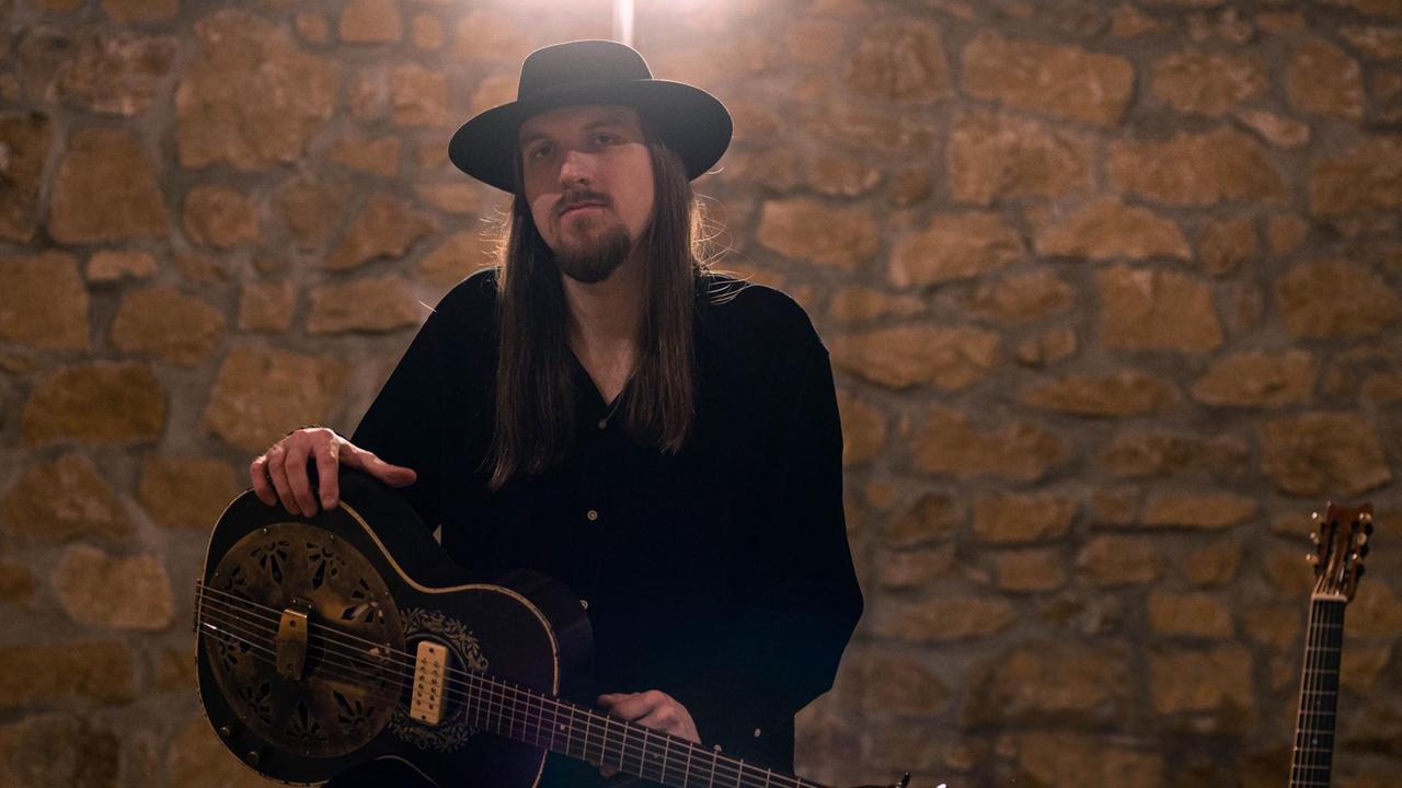 Ein Mann mit langen Haaren und Hut sitzt auf einem Hocker. Er hält eine Gitarre in den Händen.
Ein Mann mit langen Haaren und Hut sitzt auf einem Hocker. Er hält eine Gitarre in den Händen.